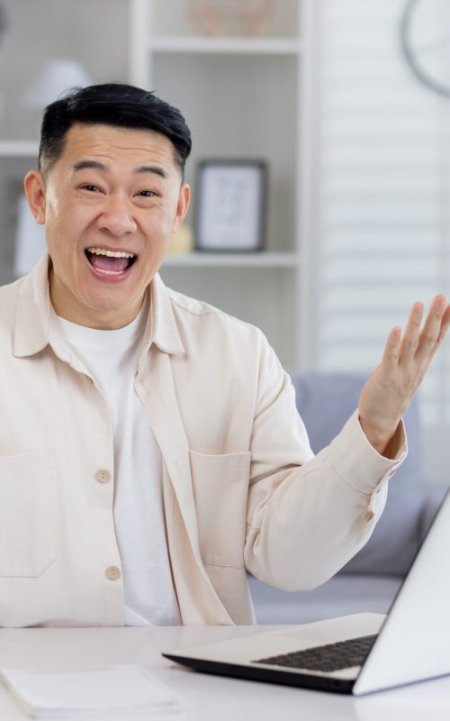 Portrait of successful Asian businessman mature man working remotely from home freelancer, living room sitting at table looking at camera and happy holding hands up celebrating victory and triump.